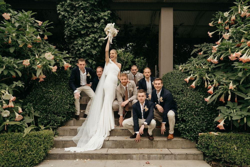 Wedding party poses playfully on garden steps surrounded by lush greenery.
