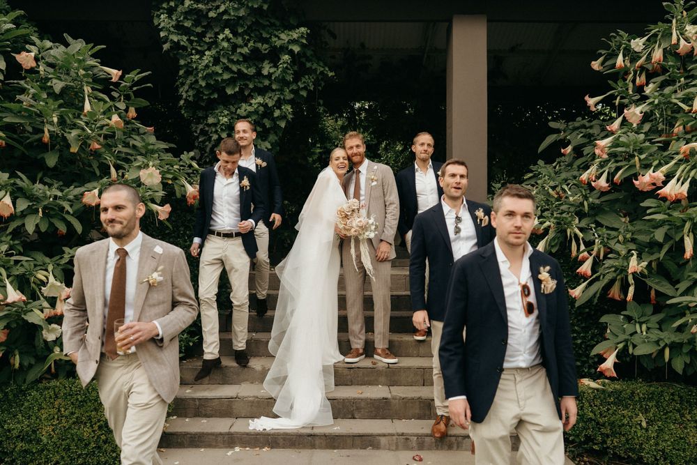 Wedding party descends stone steps between lush greenery in coordinated beige and navy attire.