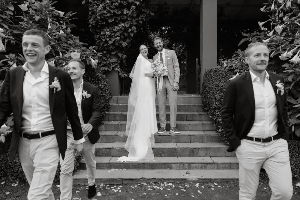 A black and white wedding portrait shows a couple on garden steps with groomsmen in the foreground.
