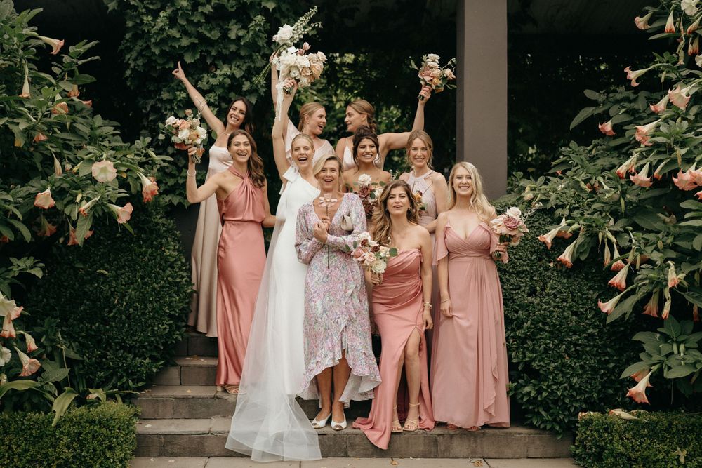 Wedding party poses on outdoor steps with bridesmaids in pink dresses surrounded by greenery.