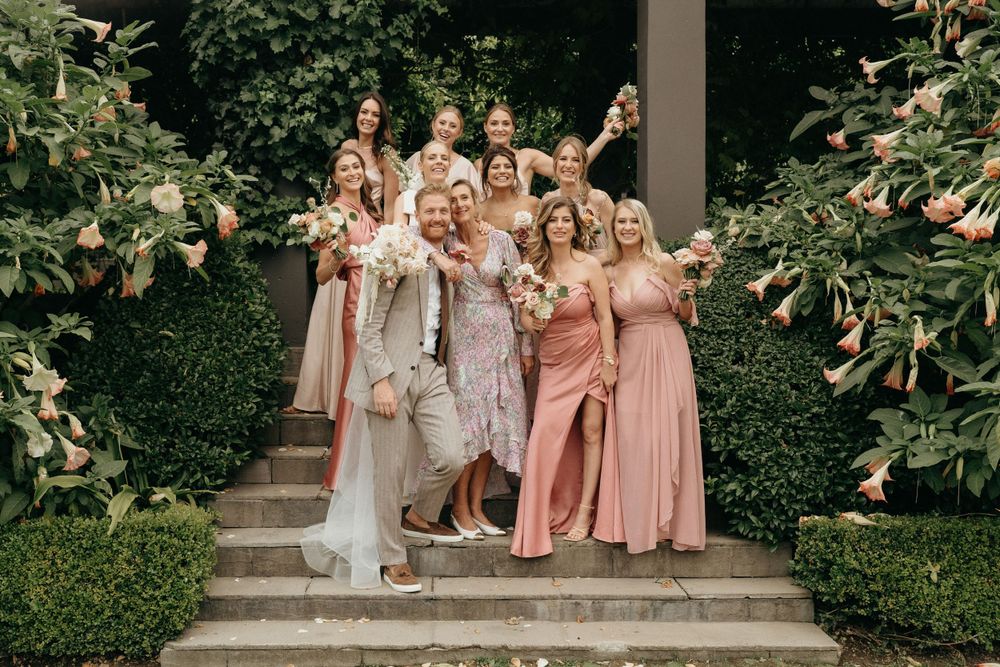 Wedding party in pink and blush dresses poses joyfully on garden steps.