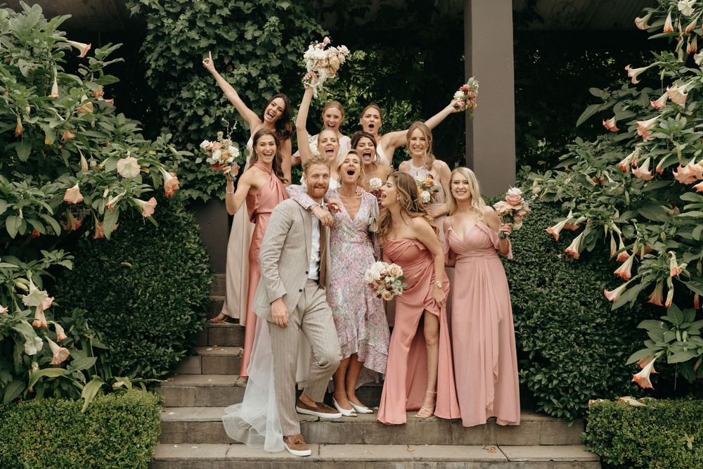 Bridal party in pink dresses celebrating joyfully on outdoor stone steps surrounded by greenery.