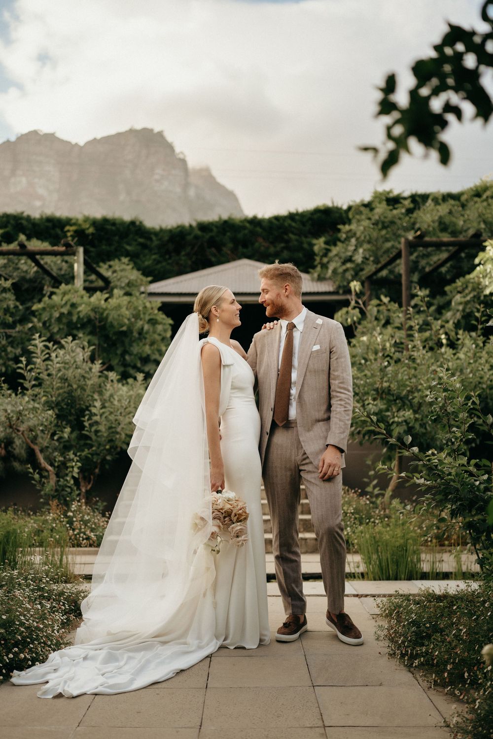 Wedding couple standing outdoors with mountain backdrop in scenic garden setting.