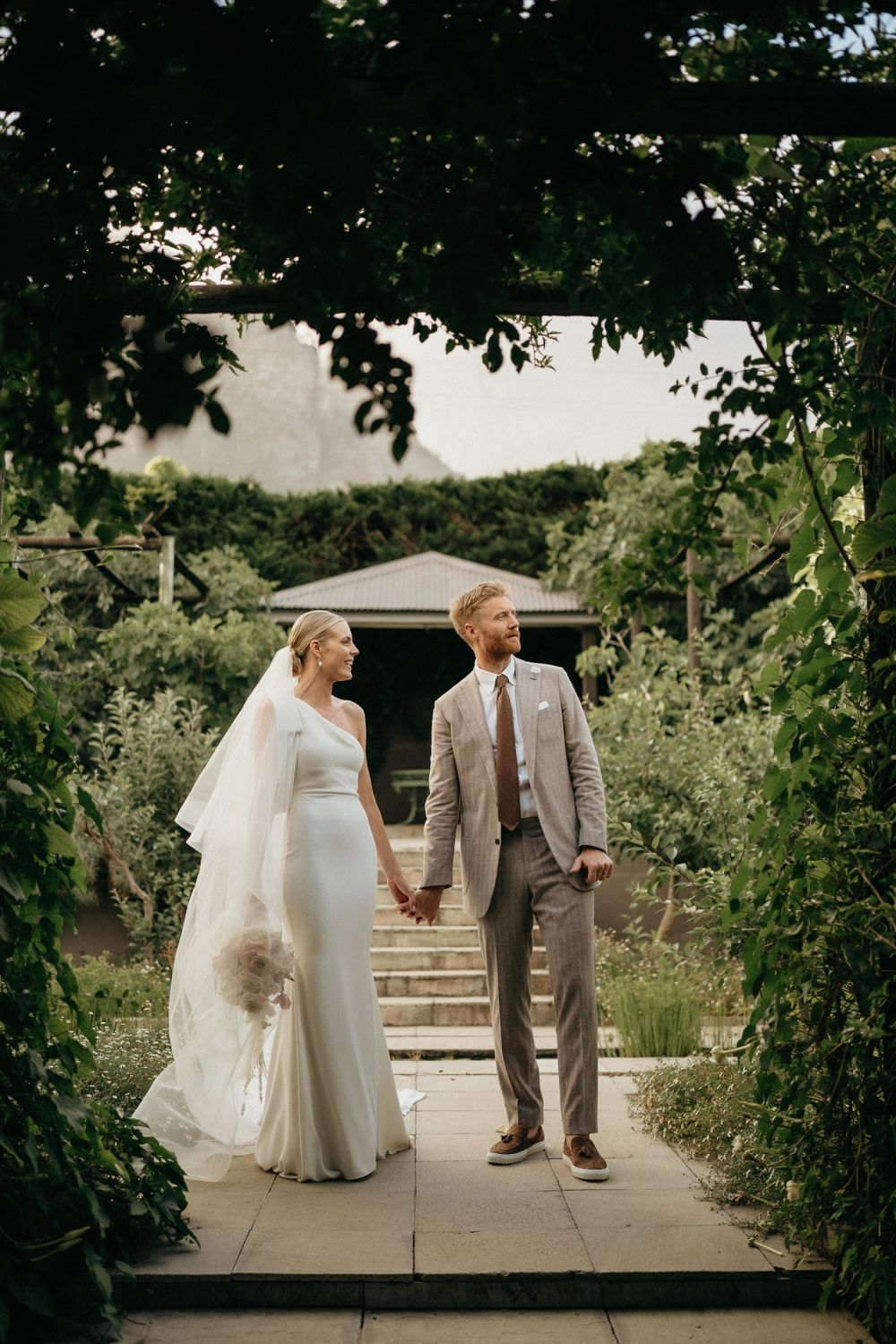 Wedding couple walking through garden archway framed by lush greenery.