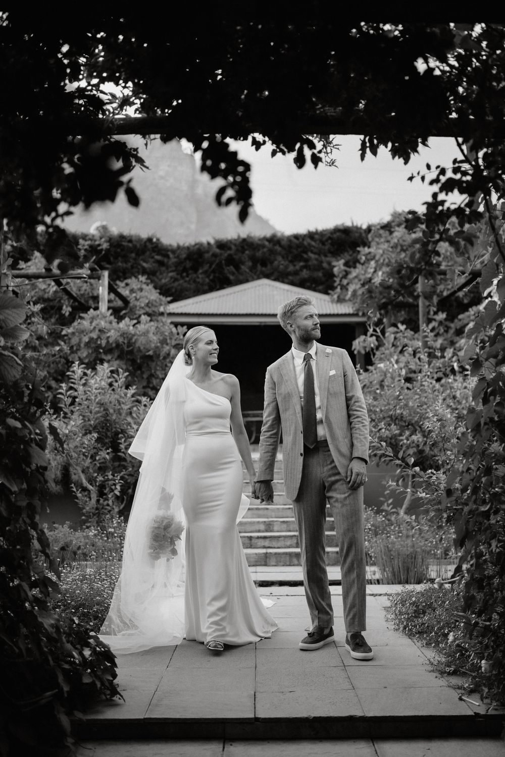 A romantic black and white wedding photo of a couple walking together on a garden path.