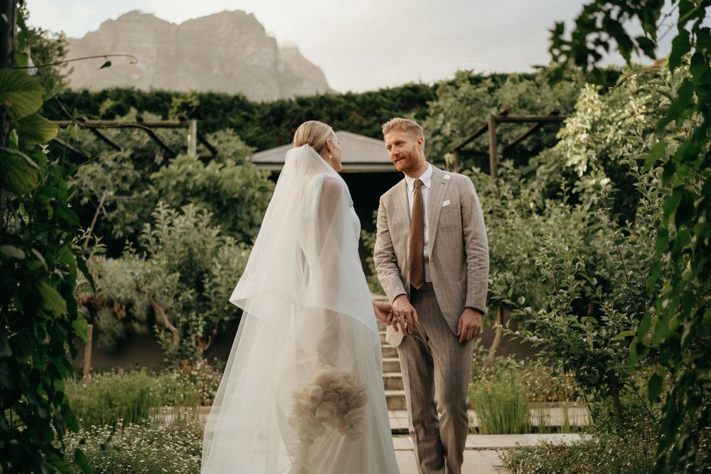 Newlyweds share intimate moment in garden with dramatic mountain backdrop.