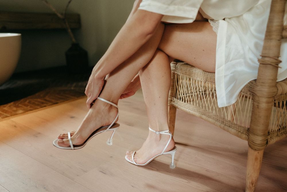 Close-up of elegant white wedding shoes being put on while seated on a rustic chair.