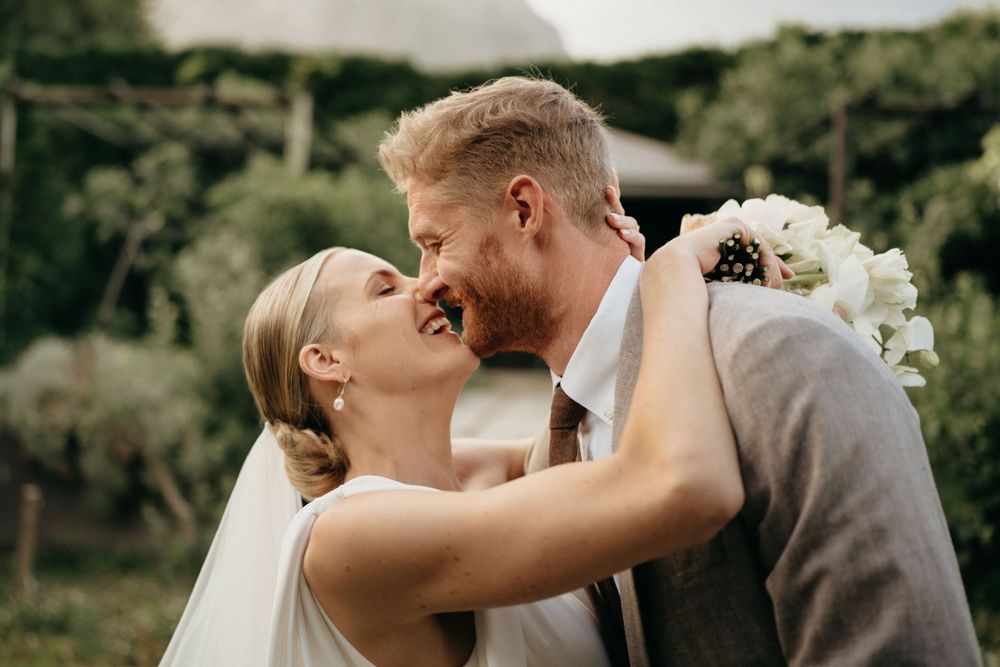 A romantic wedding couple shares an intimate kiss in an outdoor setting with white flowers and greenery.