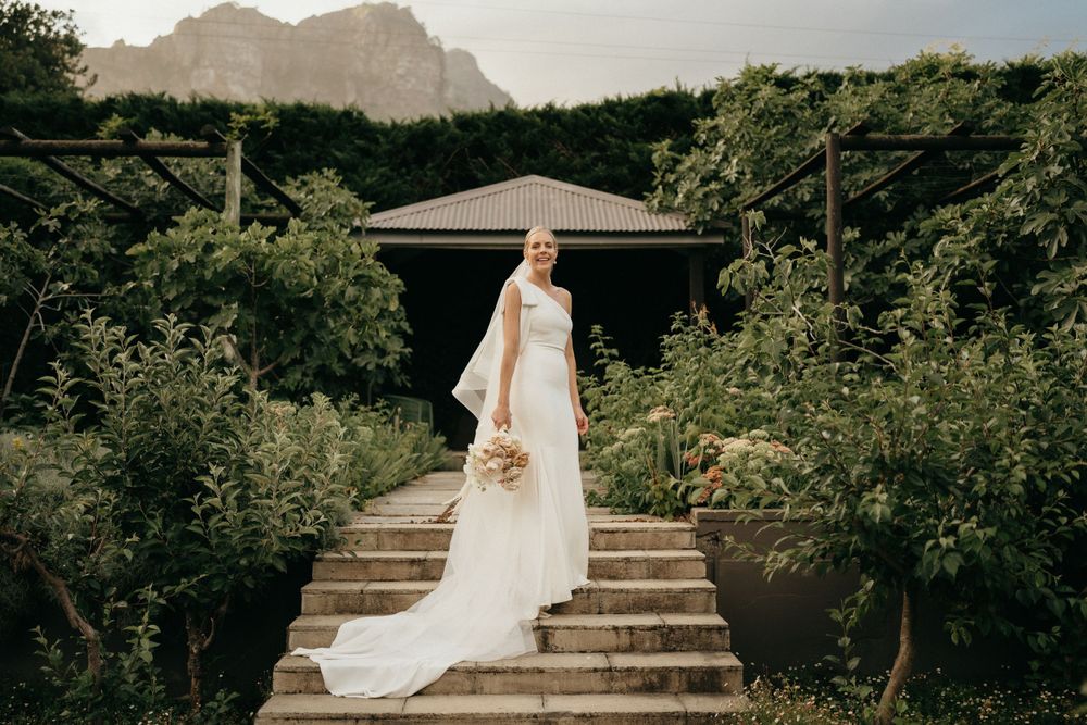 A scenic wedding photo at a garden venue with stone steps, lush greenery, and mountains in the background.