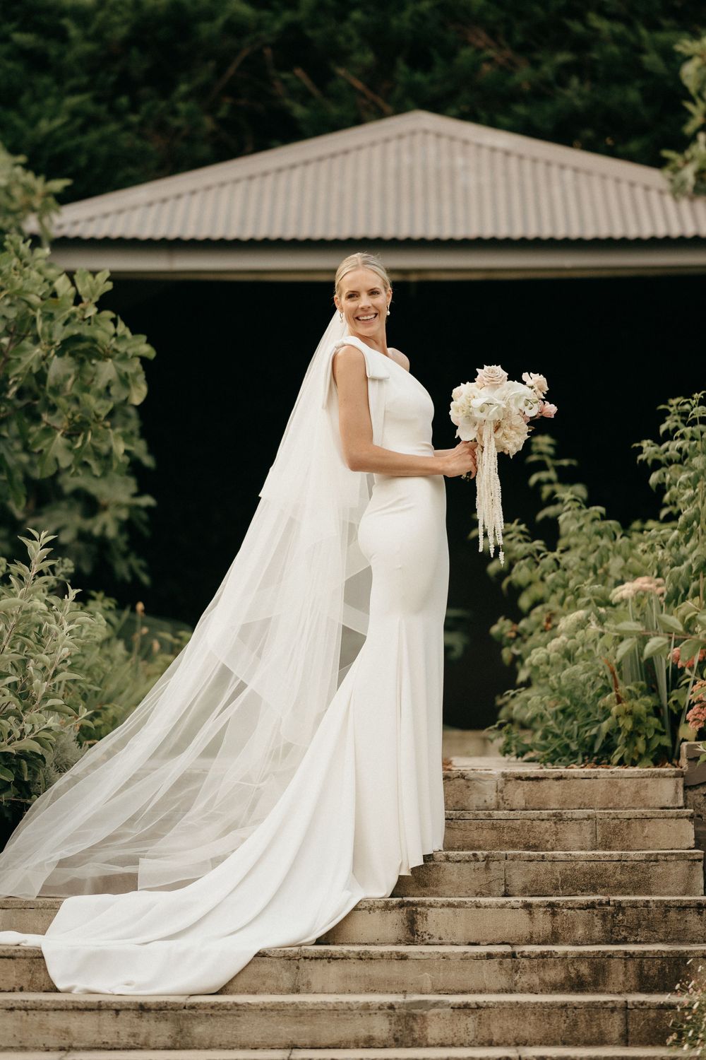 An elegant bride in a fitted white wedding gown with long flowing veil poses on stone steps in a garden setting.