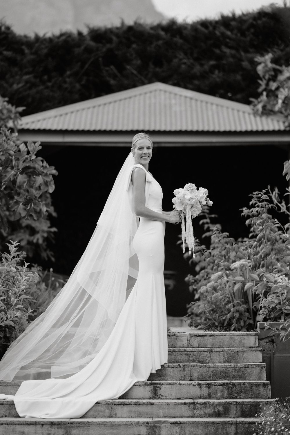 An elegant bride in a form-fitting white dress and long veil poses on stone steps in a garden setting.