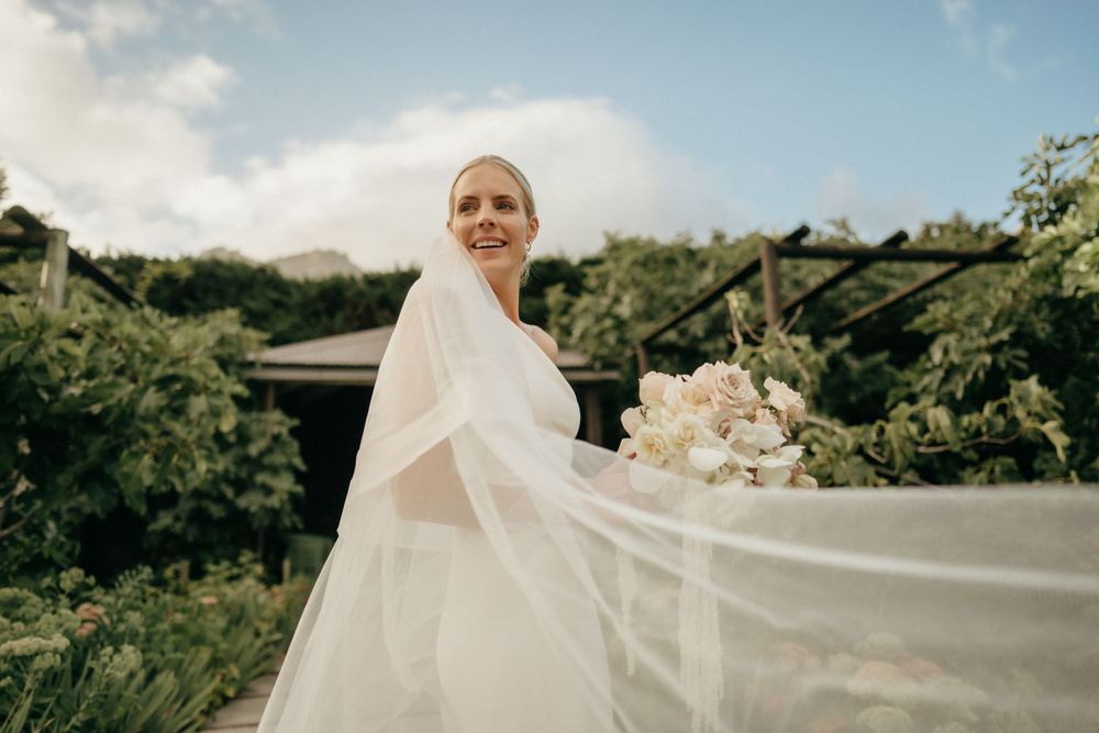 A radiant bride in a flowing white veil holds a bouquet against a lush green outdoor landscape.
