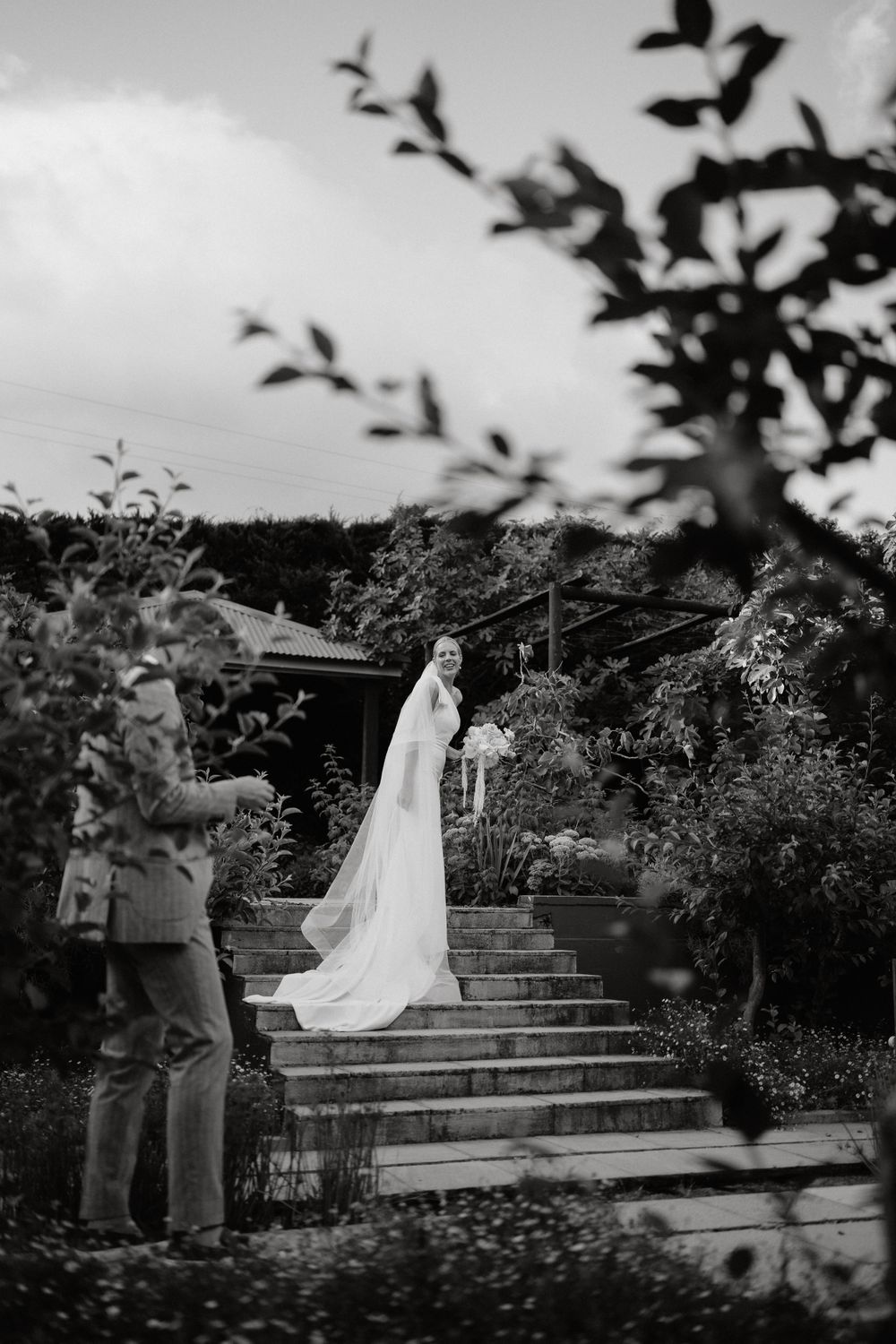 Black and white photo of a bride in flowing veil descending garden stairs.