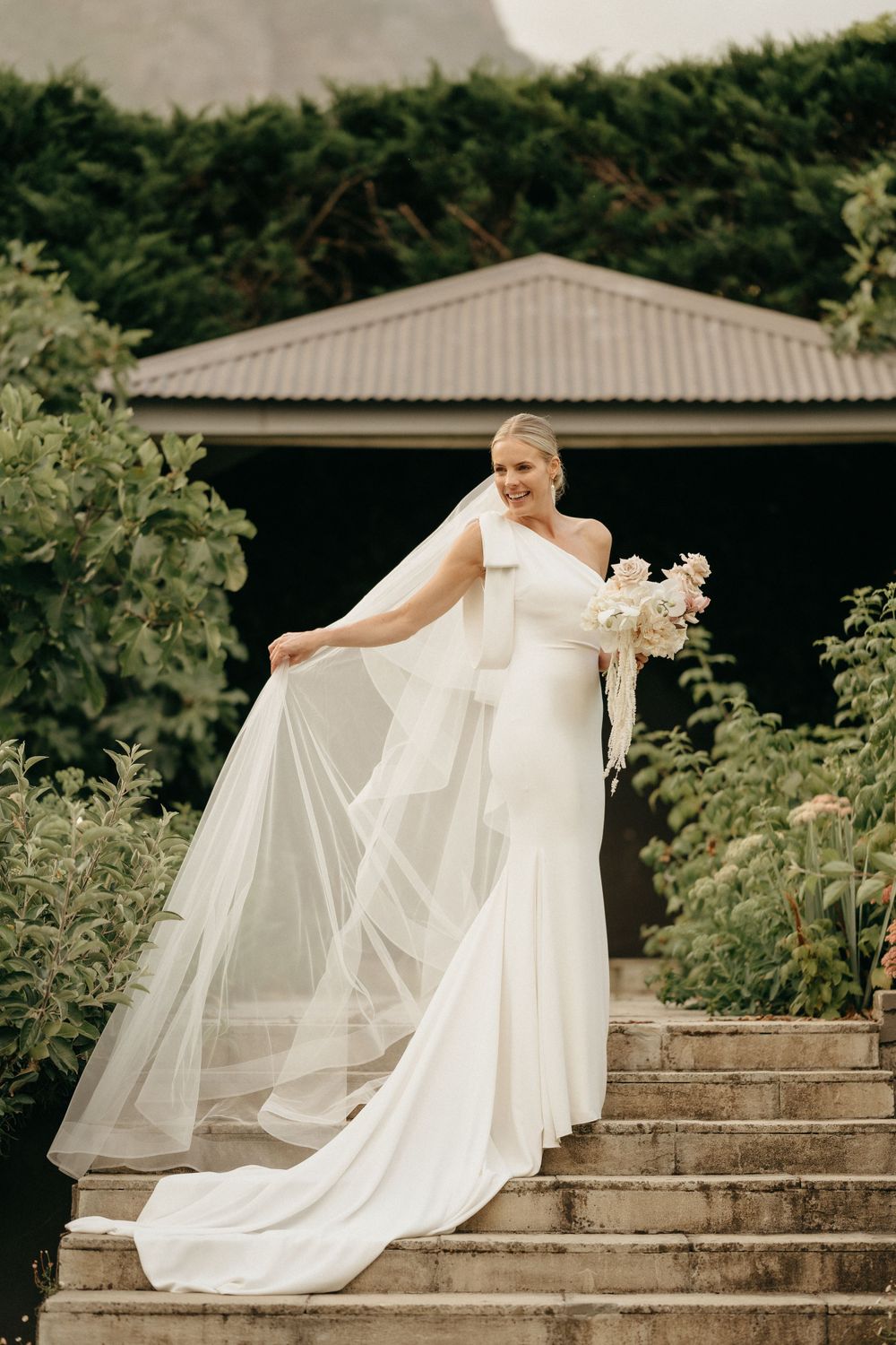 A bride in a flowing white dress stands gracefully on stone steps surrounded by greenery.
