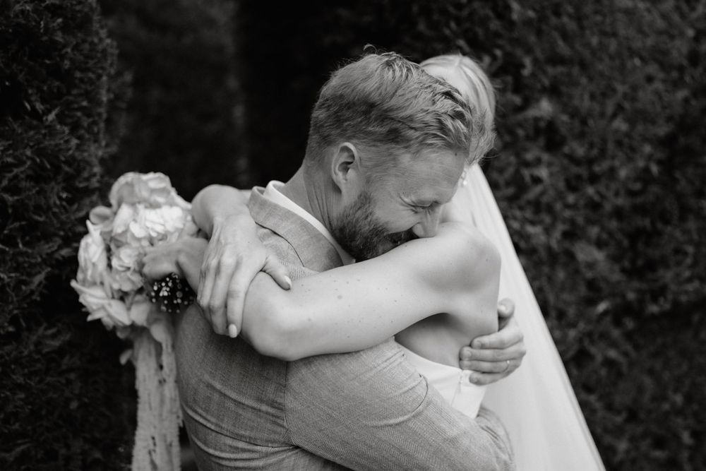 A black and white photograph shows a tender embrace between newlyweds against a dark background.
