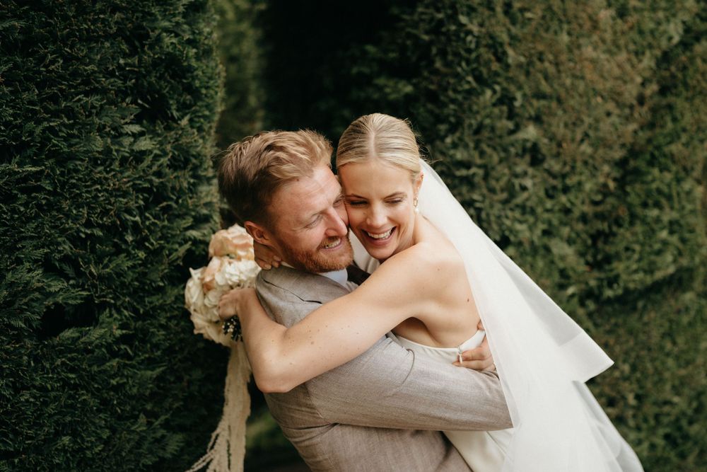 Joyful couple embraces and laughs together in a garden setting with green hedge backdrop.