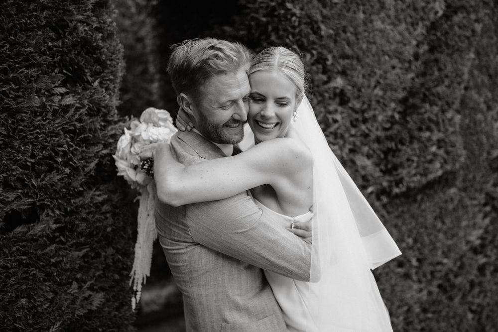 Black and white photo of couple embracing during wedding celebration in garden setting.