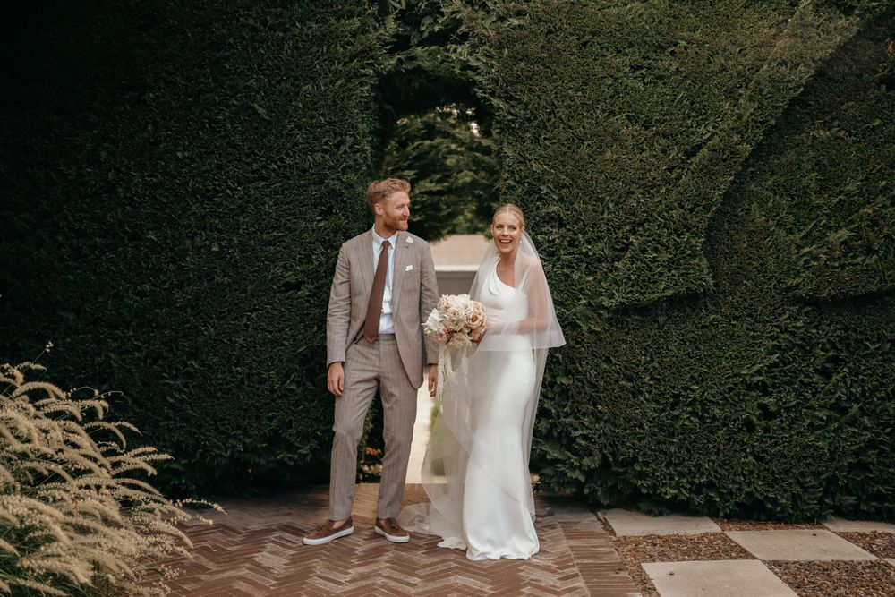 A couple poses together during their wedding ceremony against a lush green hedge wall backdrop.