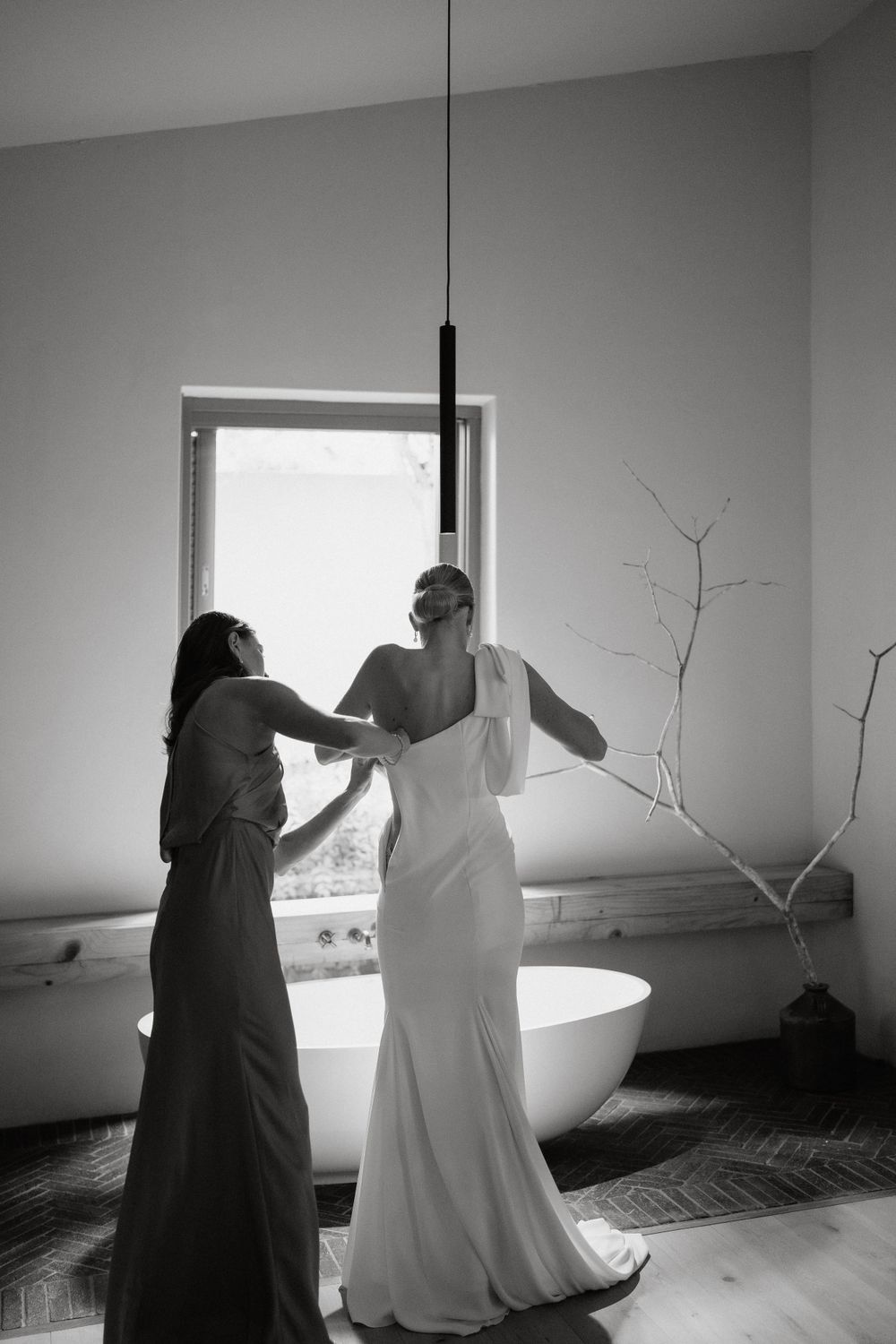 Black and white photo of a bride getting dressed in an elegant white gown next to a window.