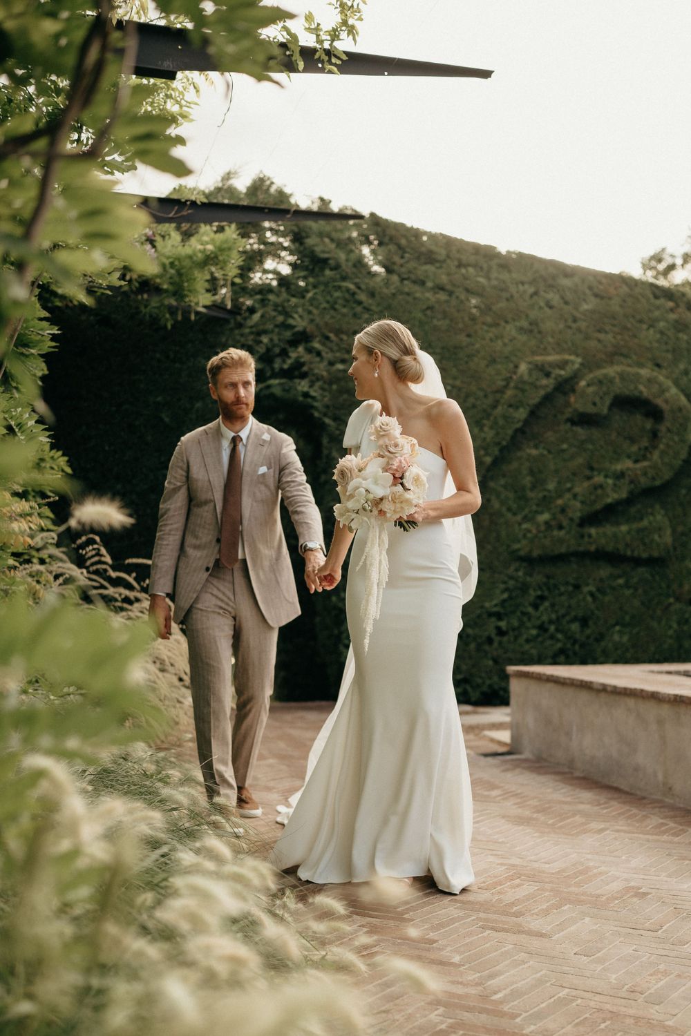 A couple on their wedding day walk together in a garden setting with greenery and stone architecture.