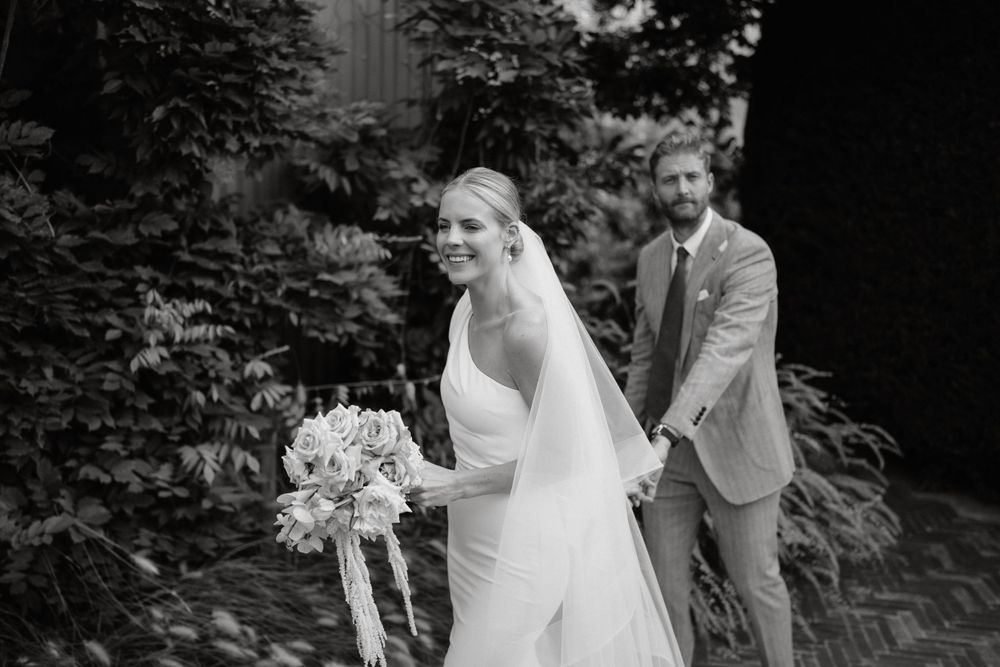 A black and white wedding portrait showing a bride holding a bouquet while walking with her groom in a garden setting.