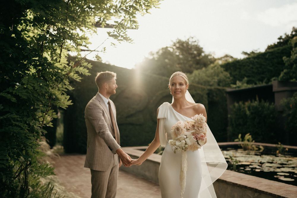 Couple stands in garden during golden hour sunset, bride in elegant one-shoulder gown with flowing bouquet.