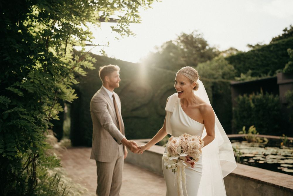 Couple holds hands in sunlit garden setting, bride wearing modern one-shoulder wedding gown.