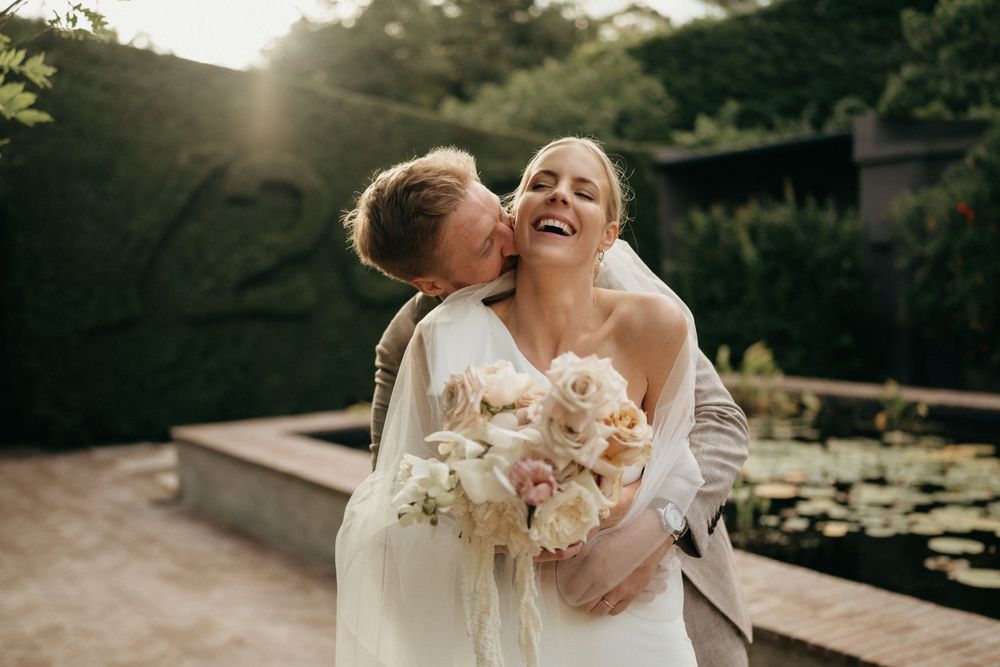 Romantic sunlit wedding moment as couple embraces with blush pink bouquet by garden pond.