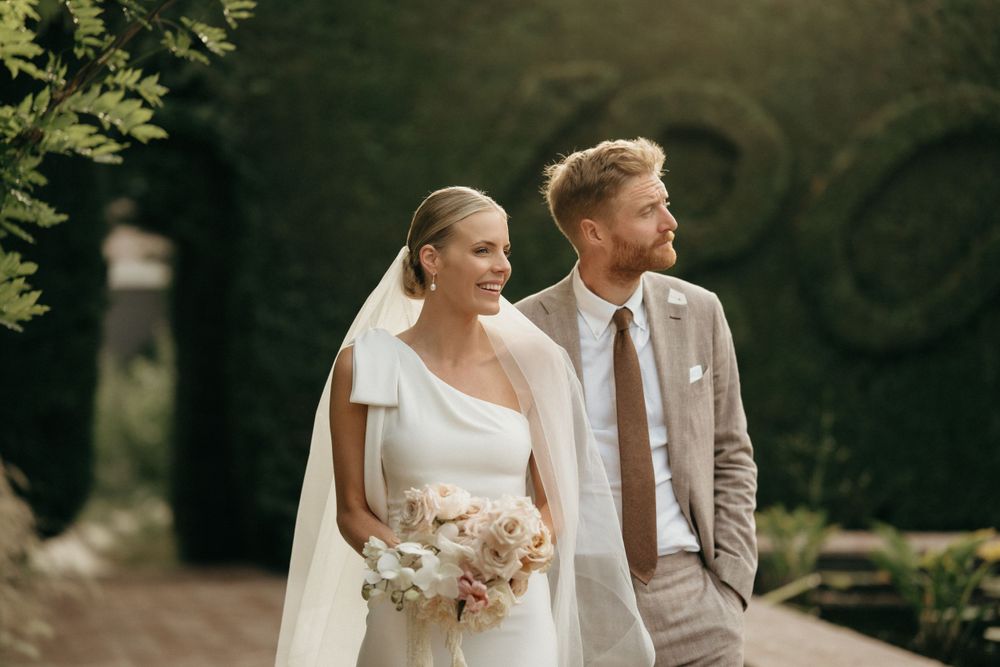 Elegant wedding couple poses in garden, bride in one-shoulder gown with flowing veil and light bouquet.