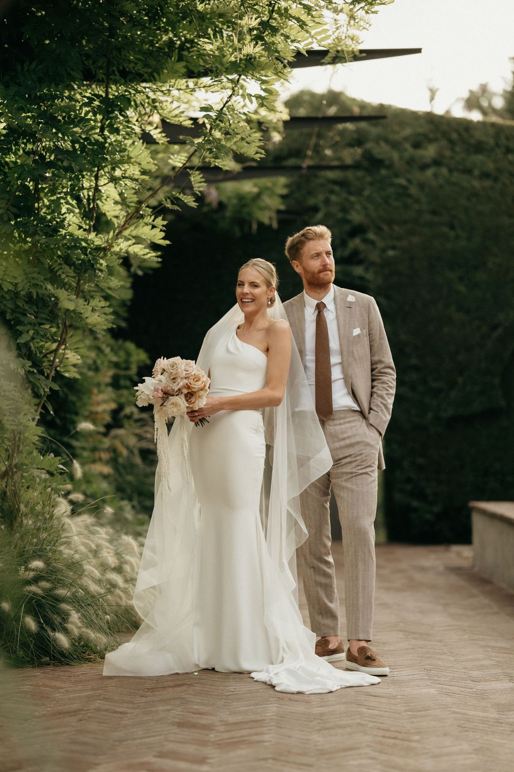 Elegant wedding couple poses in a garden setting with flowing white dress and tan suit.