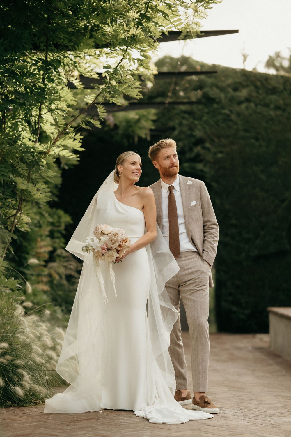 Wedding couple poses outdoors in elegant attire with bride in white gown and groom in beige suit at garden venue.