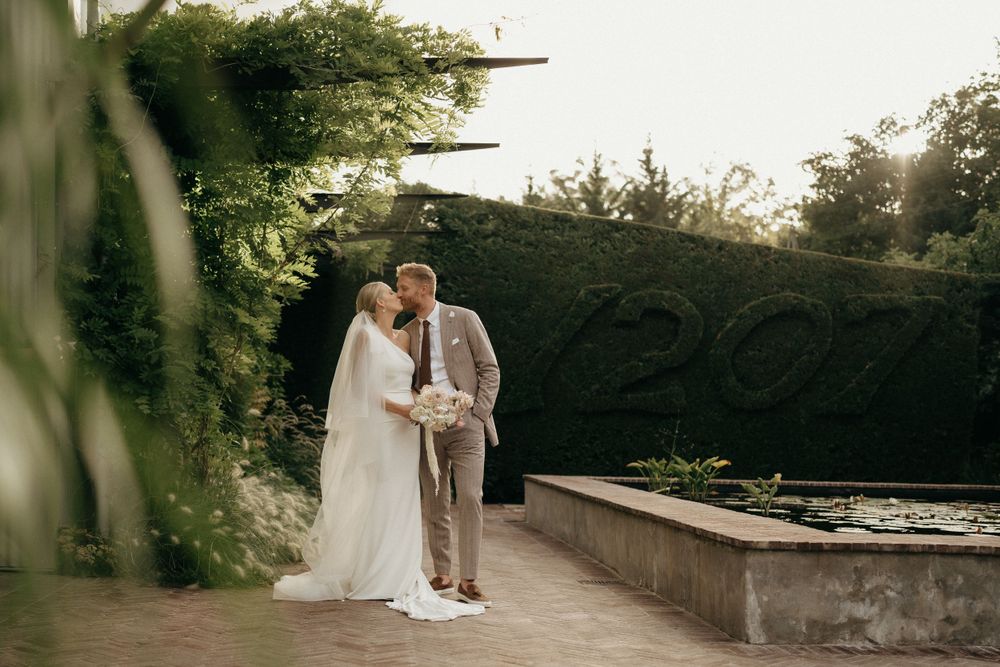 A couple in wedding attire embraces on a garden terrace during sunset.