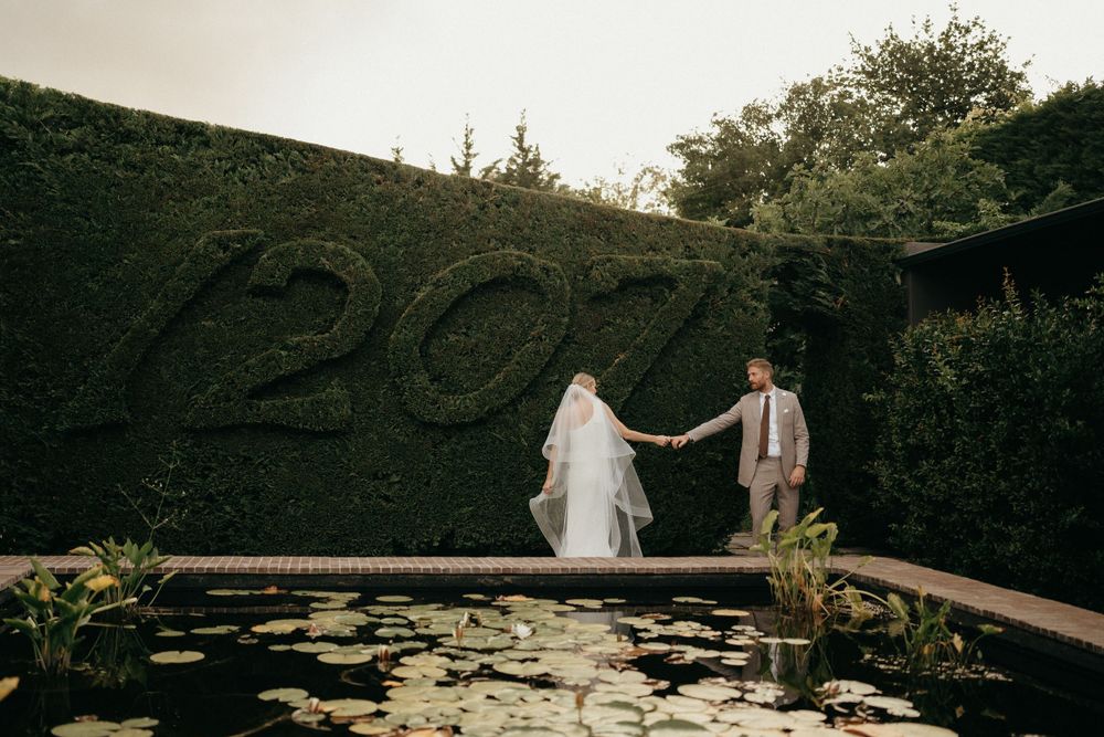 Couple standing near pond with '20' number design carved into garden hedge wall.