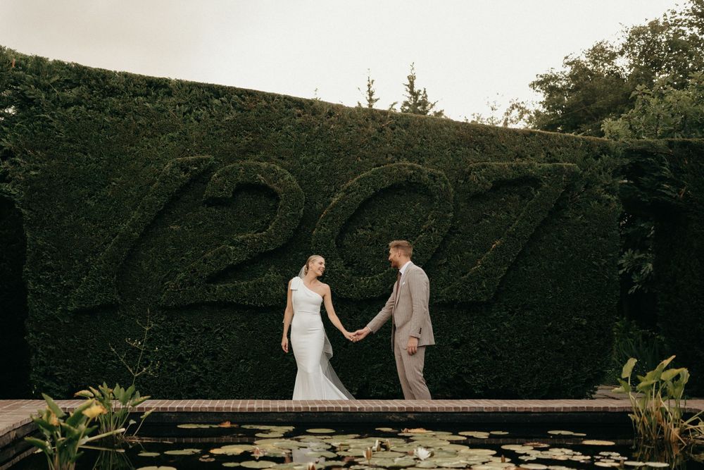 A couple stands near a decorative hedge wall displaying the year 1927 in a garden setting.