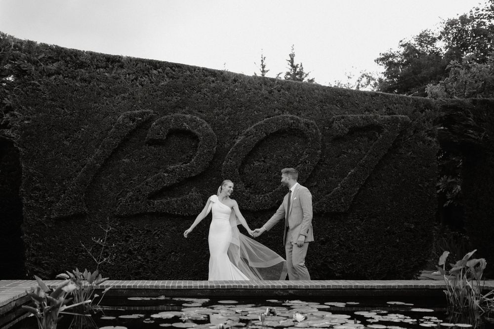 Black and white photo of couple posing by a pond with decorative hedge numbers in the background.