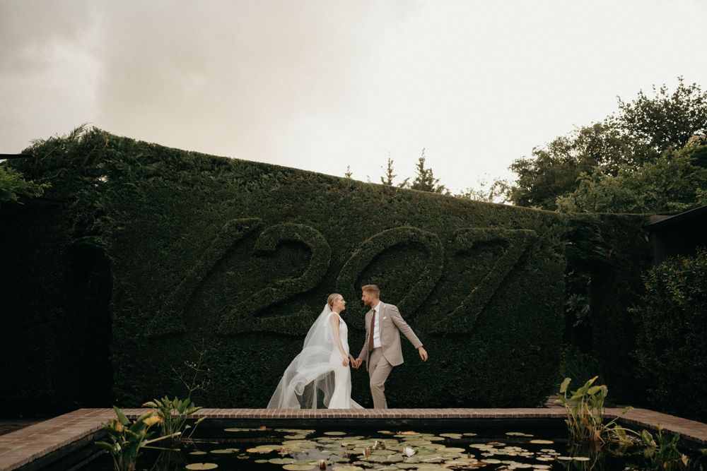 Couple walking together on a bridge against a backdrop of lush green hedges.