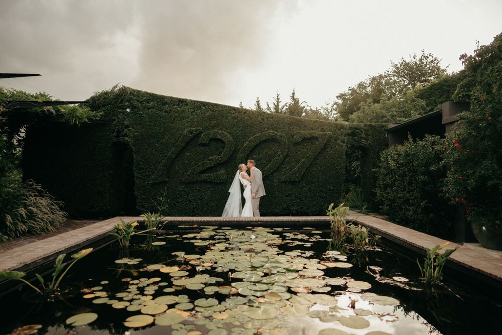 Romantic wedding couple silhouetted against moody sky at garden pond with lily pads.