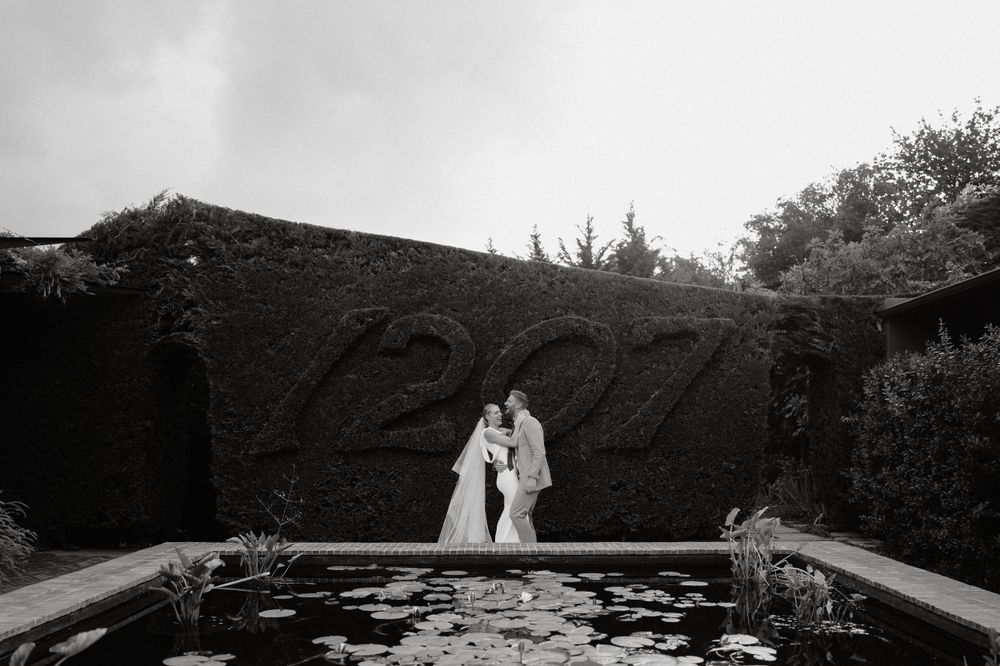 A dramatic black and white photo of a couple by a reflective pond with topiary hedges.