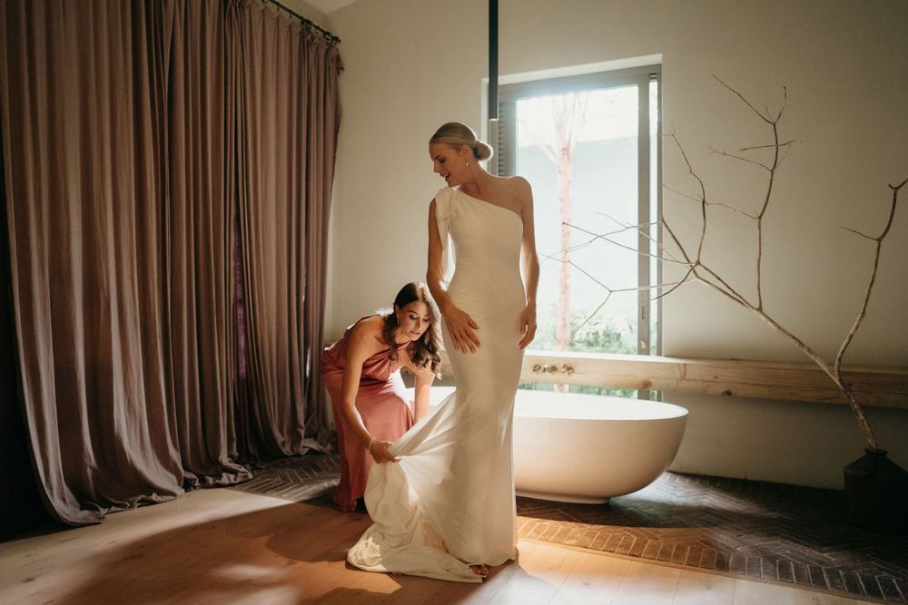A wedding dress being fitted and adjusted in a sunlit room with flowing curtains and hardwood floors.