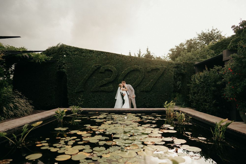 A couple embraces by a tranquil pond with lily pads in a romantic garden setting.