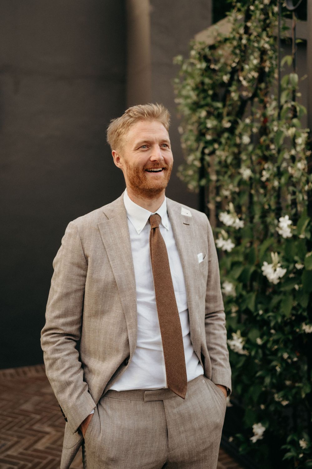 A person in a light gray suit with brown tie stands smiling against a wall with climbing vines.