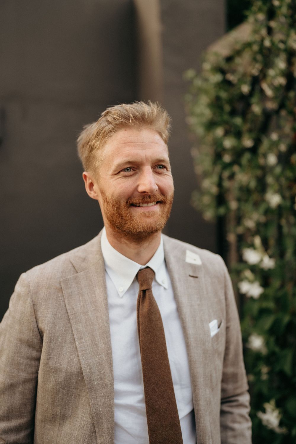 A person wearing a light gray suit with a brown tie smiles warmly while standing outdoors near greenery.