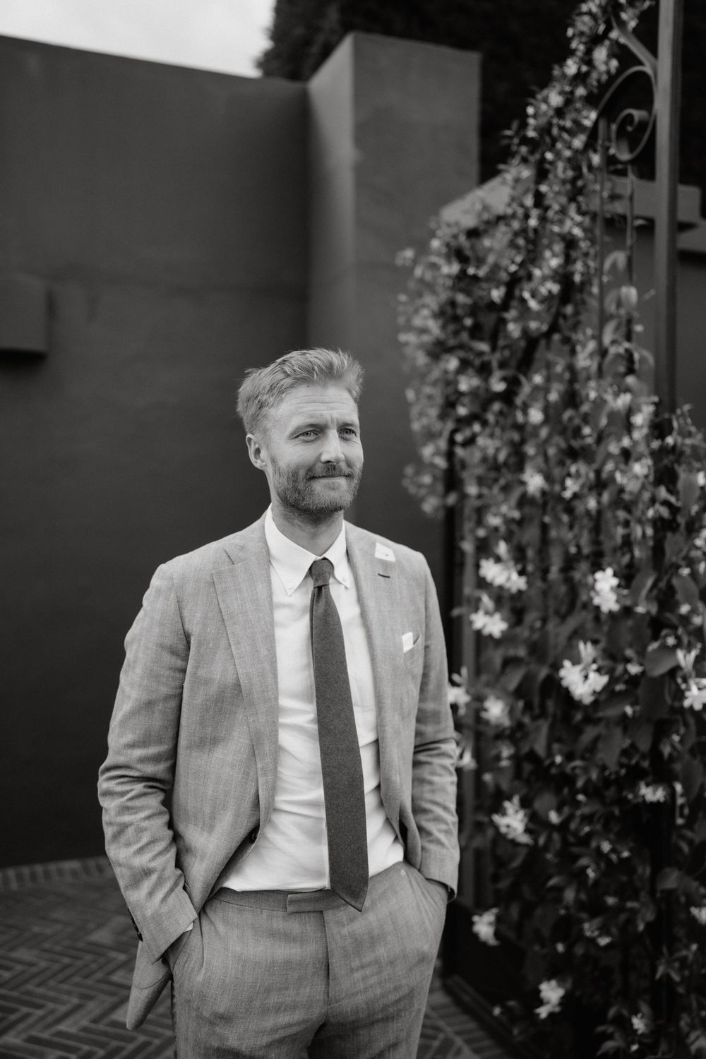 Person in grey suit standing against dark wall with flowering vines.