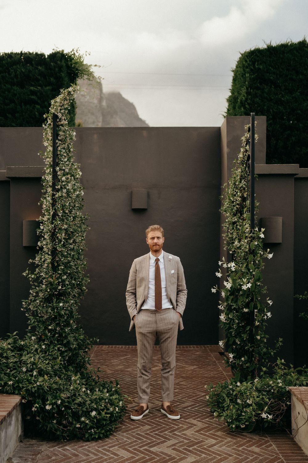 A figure in a light gray suit stands between two climbing ivy columns against a dark wall in an elegant garden setting.