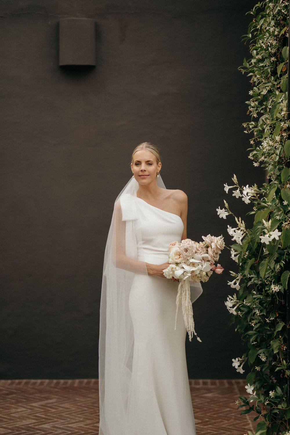 Person in elegant one-shoulder white wedding gown with veil poses against dark wall with climbing flowers.