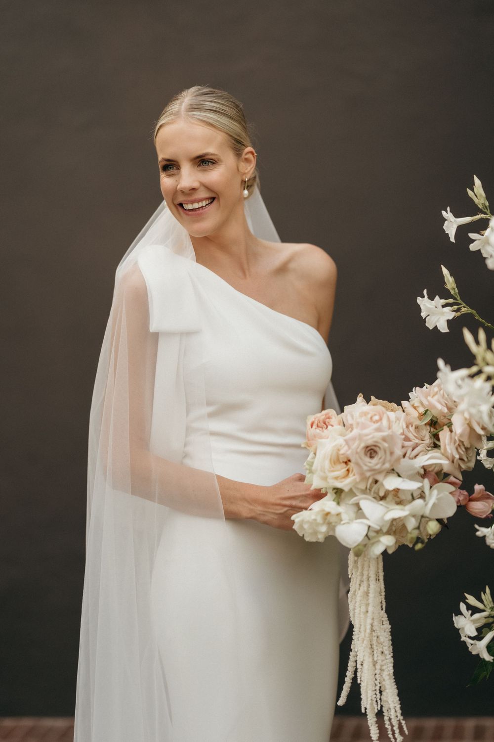 Elegant bride in one-shoulder white gown holds romantic blush and white floral bouquet with flowing veil.