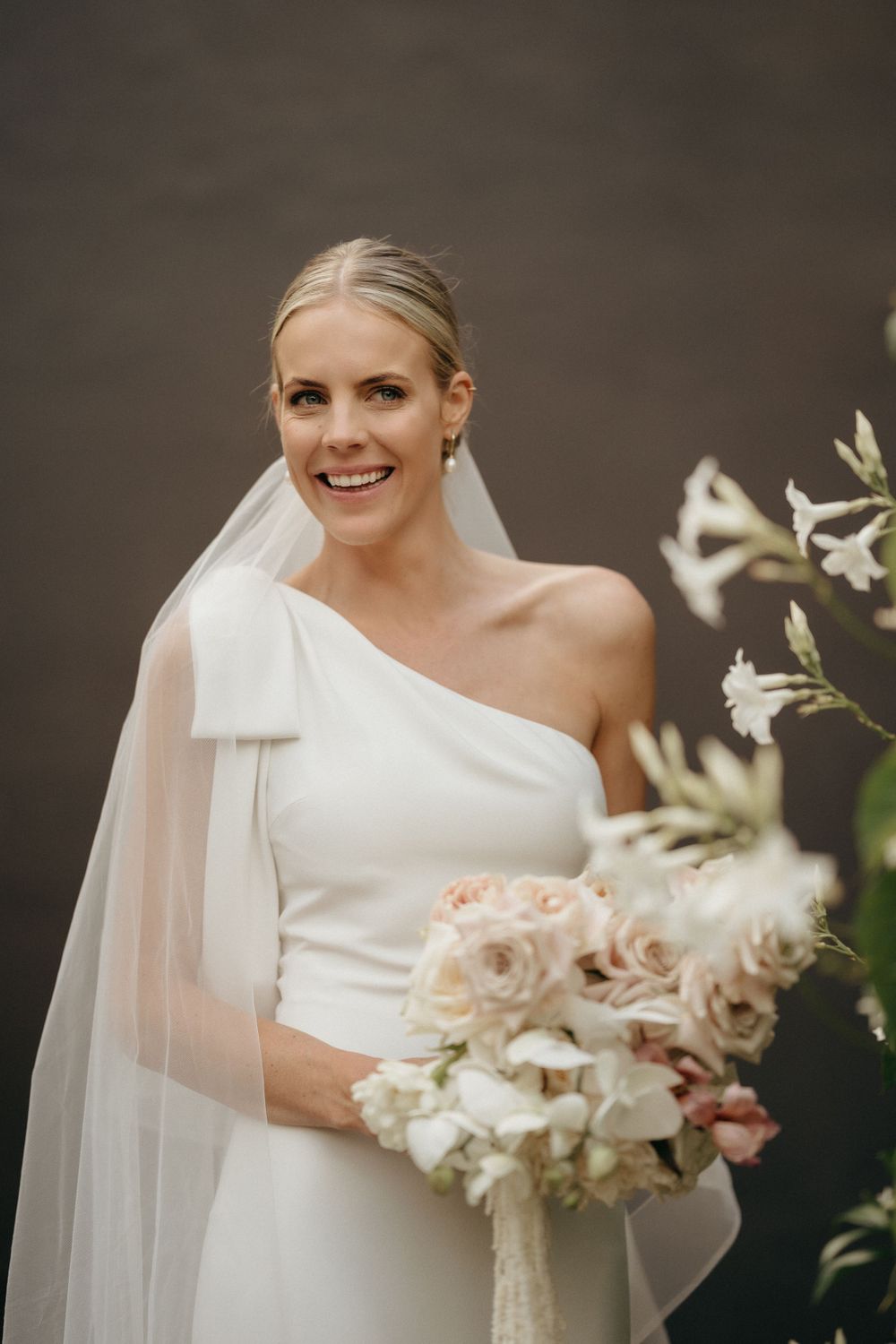 Elegant bride in one-shoulder white gown holds romantic blush and ivory bouquet.