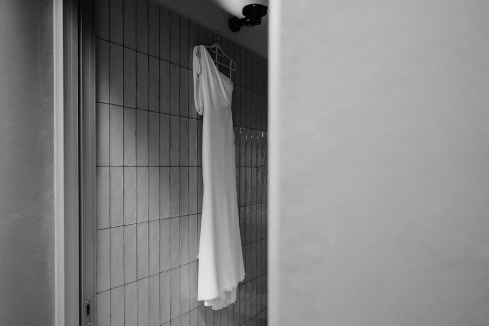 A minimalist black and white photo of a wedding dress hanging in a tiled bathroom.