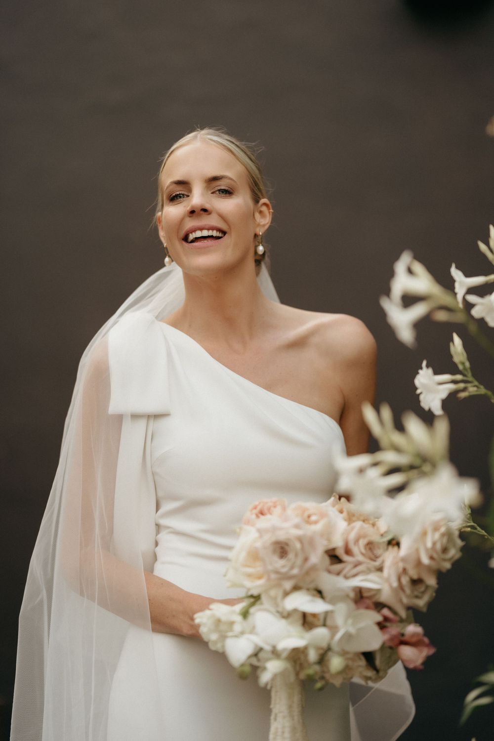 Elegant one-shoulder wedding gown with flowing veil and peachy floral bouquet.
