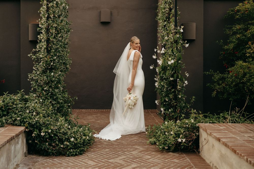 A bride in a flowing white gown and veil stands between two ivy-covered pillars on a brick pathway.